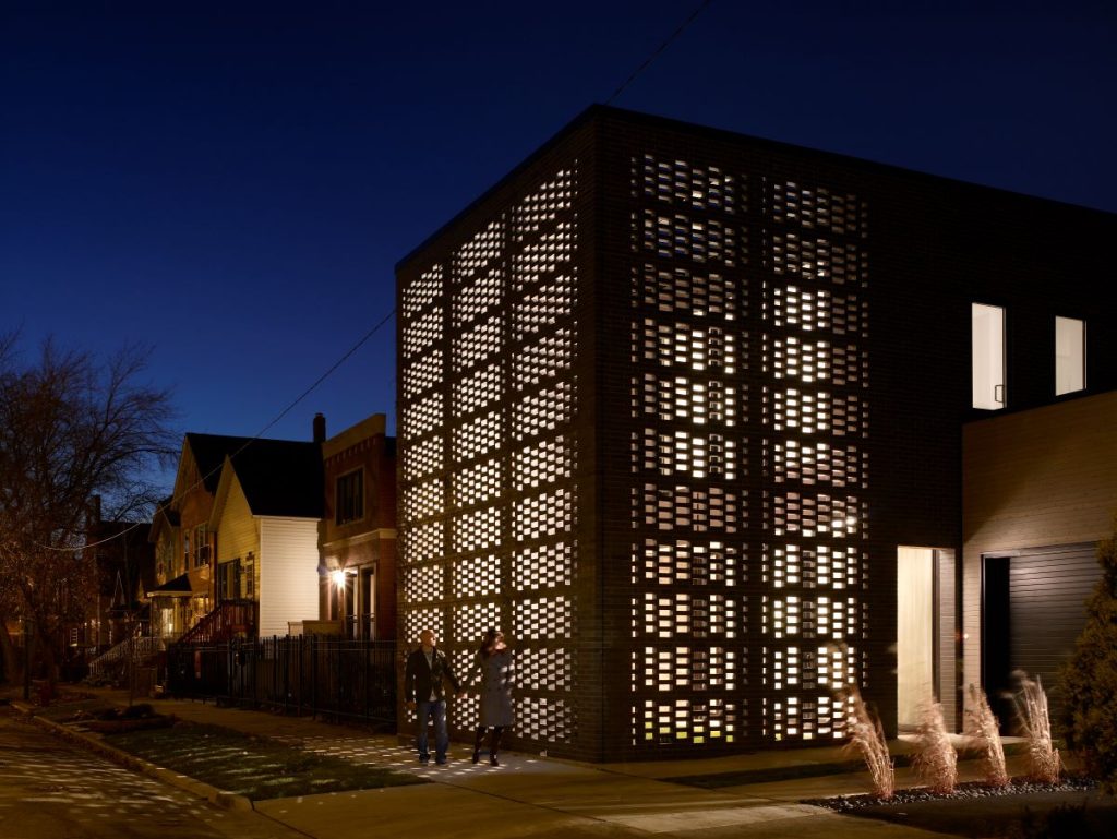 A photo of a lit up Chicago house at dusk. The brickwork facing the street suggests weaving and the wall sits flush against the sidewalk outside.