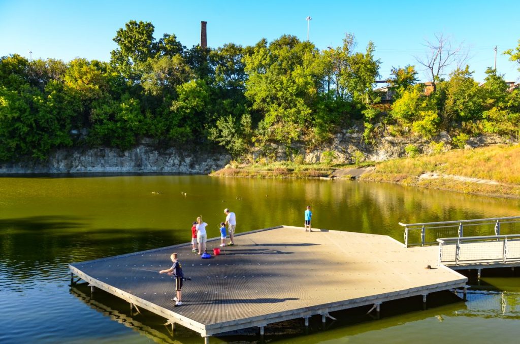 An irregular five side platform juts onto a pond at Palmisano Park. A family of six is fishing off of the platform on a sunny summer day.