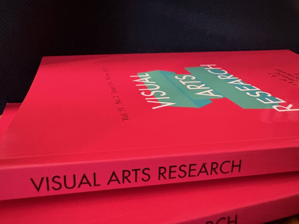 Visual Arts Research, Volume 51, No. 2, Winter 2025 with a pink background with list of editors in black text, three teal rectangular prisms behind title, stacked atop Vol. 51, No. 1, horizontally with the spines showing, in front of a black background.