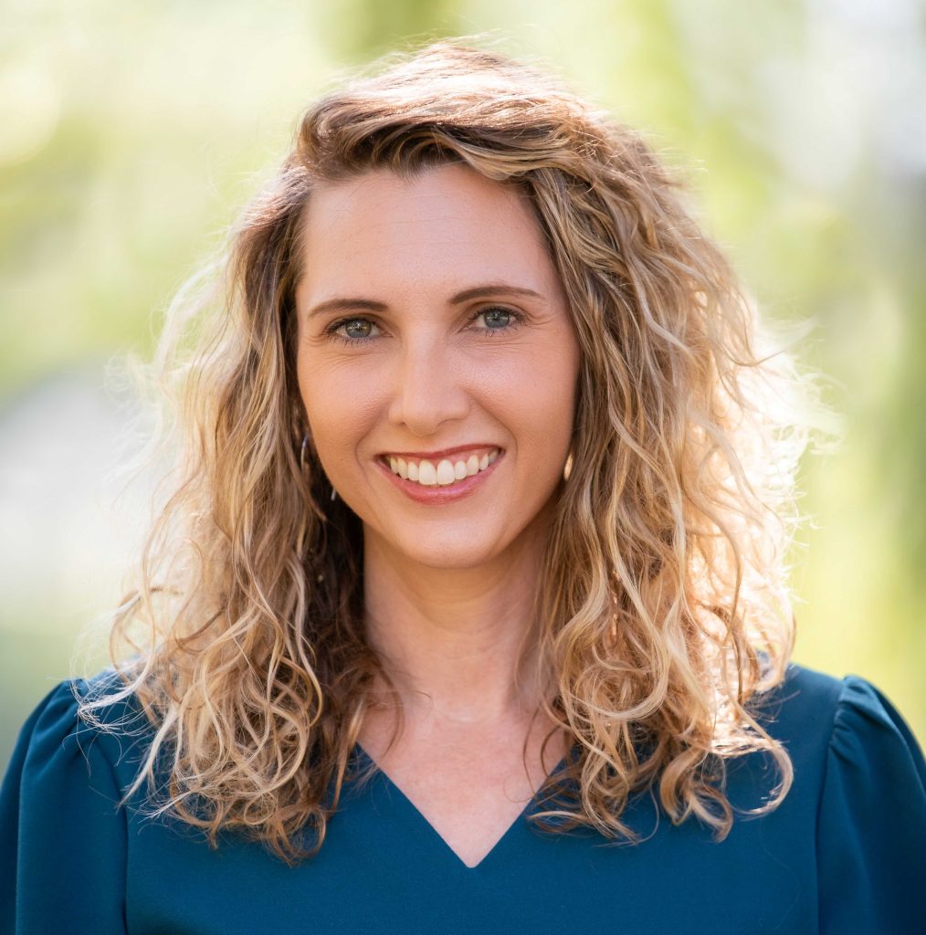 Photo of Caroline Kline, with a blurred outdoor background. Kline is wearing a blue shirt and has blond curly hair.
