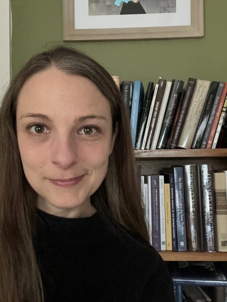 Photo of Margaret Olsen Hemming, with a bookcase background. She is wearing a black shirt and has long straight brown hair.
