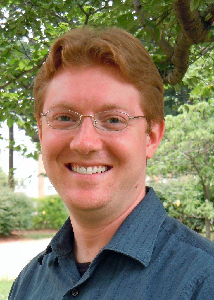 Photo of Christian McWhirter wearing a collared shirt and glasses, smiling, in front of a background of green trees and shrubs.