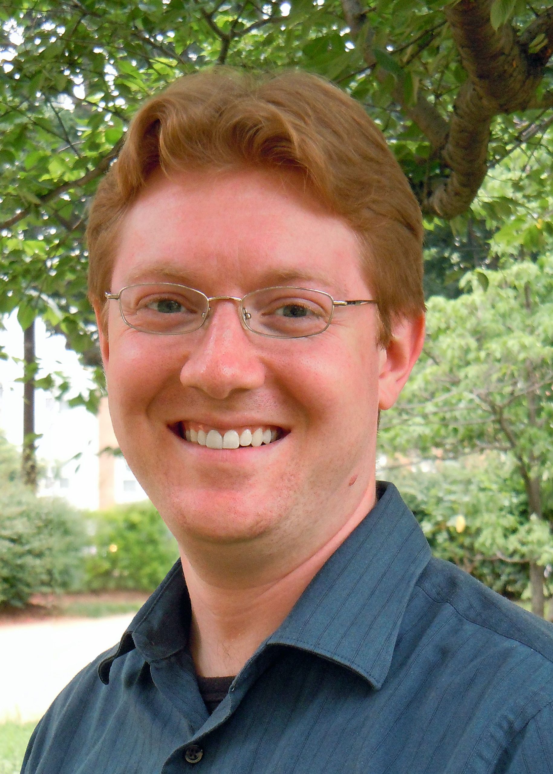 Photo of Christian McWhirter wearing a collared shirt and glasses, smiling, in front of a background of green trees and shrubs.
