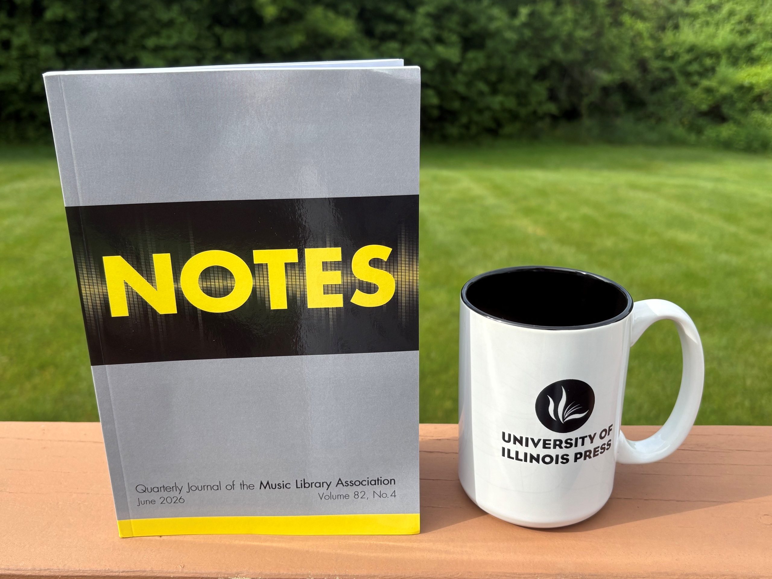 Alt text: Copy of Notes, with a gray and yellow cover graphic imitating sound waves, standing next to a University of Illinois Press mug on an outdoor table with a grass lawn in the background.