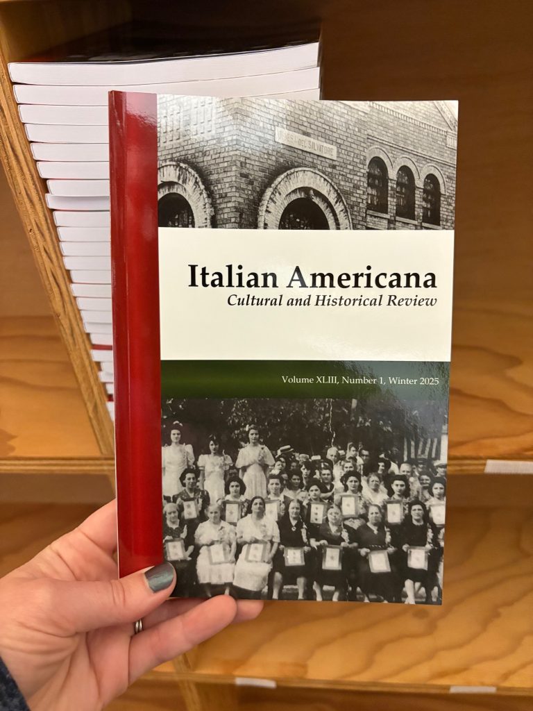 An issue of Italian Americana being held up in front of bookcase with a stack of journals.