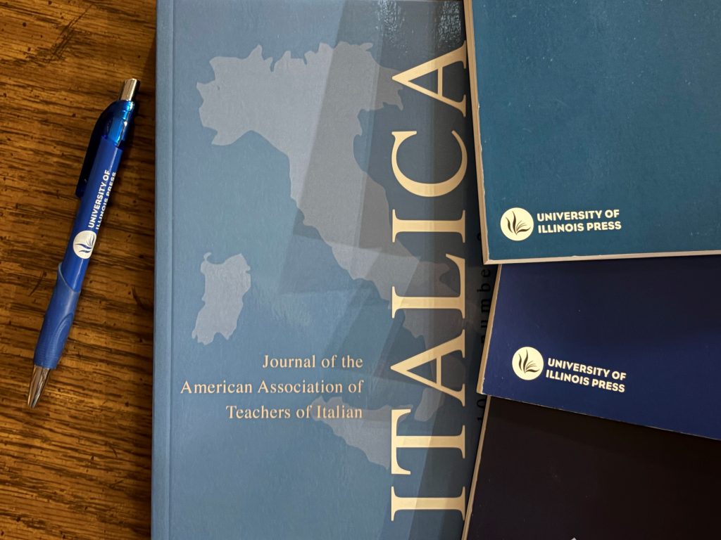 Copy of Italica laying on a wooden table next to a University of Illinois Press-branded pen and other blue journals