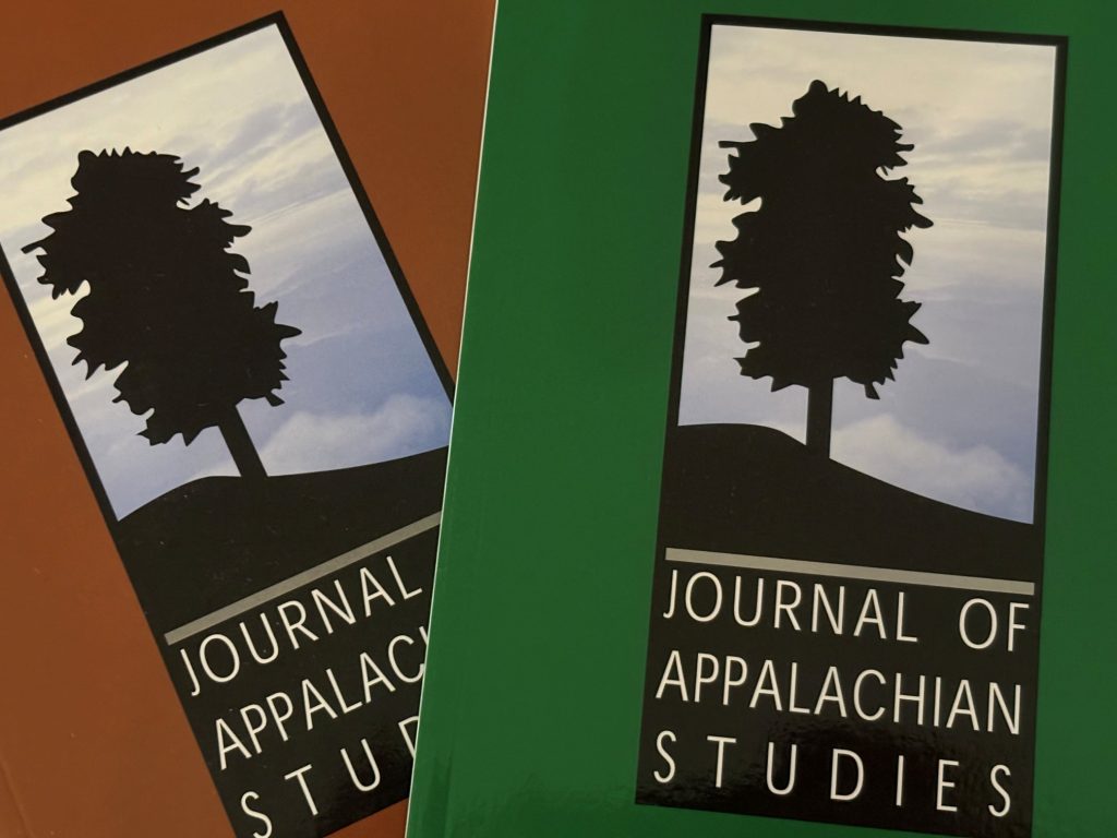 Two issues of Journal of Appalachian Studies, one with a brown cover and one with a green cover each with a silhouette of a tree on a hill in front of a cloudy sky, blue background